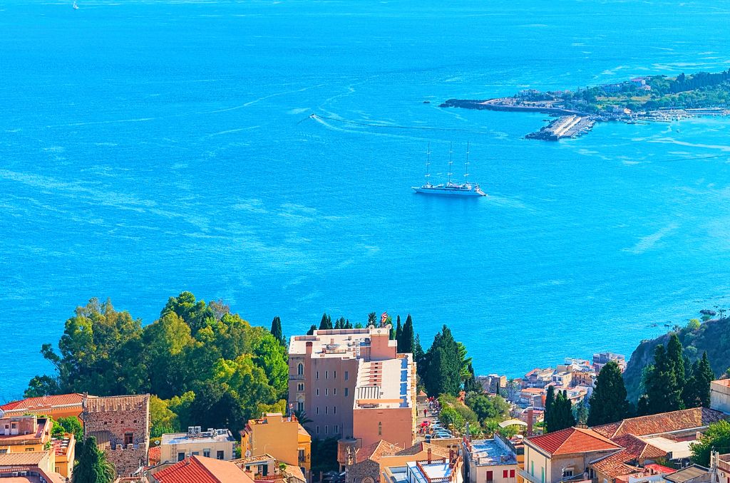 Blick von oben auf eine Küstenstadt mit rötlichen Dächern. Dahinter erstreckt sich ein azurblaues Meer, auf dem ein großer Segler ankert. Blick von oben auf eine Küstenstadt mit rötlichen Dächern. Dahinter erstreckt sich ein azurblaues Meer, auf dem ein großer Segler ankert.