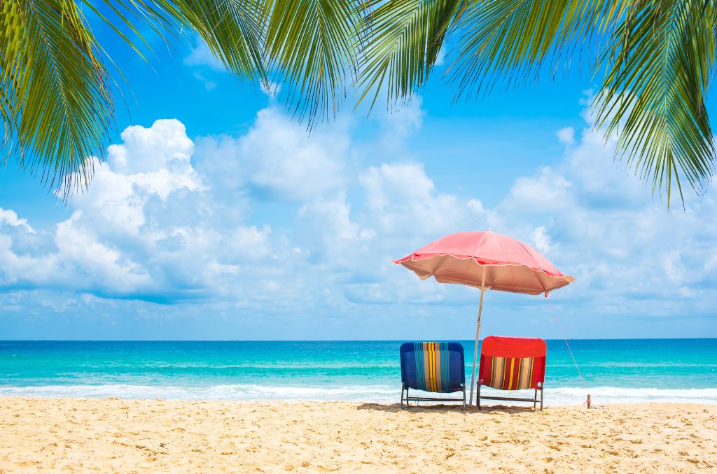 Zwei gestreifte Liegestühle unter einem roten Sonnenschirm an einem Sandstrand mit Blick auf das türkisfarbene Meer. Palmblätter rahmen das Bild oben ein. Zwei gestreifte Liegestühle unter einem roten Sonnenschirm an einem Sandstrand mit Blick auf das türkisfarbene Meer. Palmblätter rahmen das Bild oben ein.